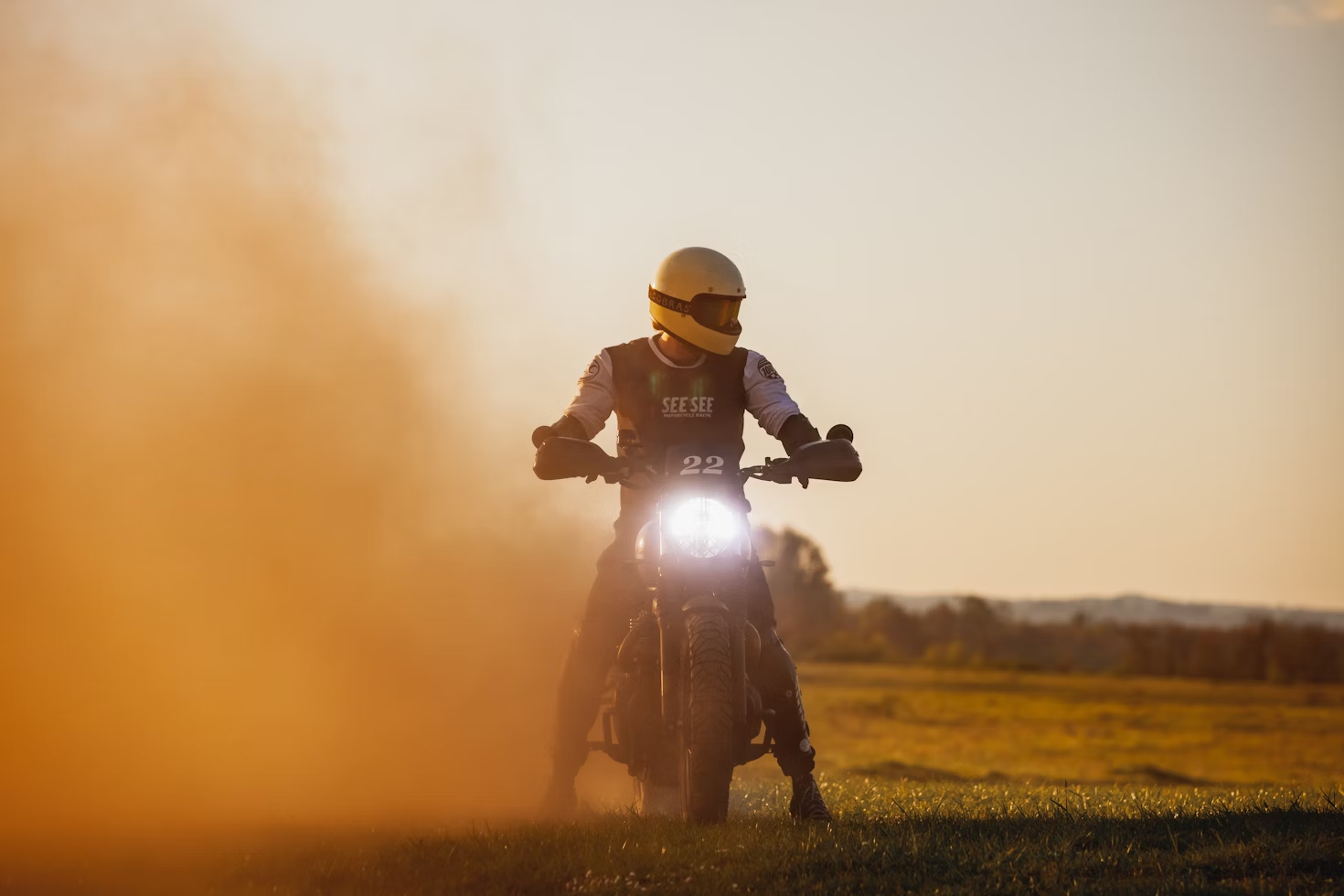 Motorcyclist wearing a vintage yellow helmet rides through a cloud of dust at sunset, headlight beaming across an open grassy field.