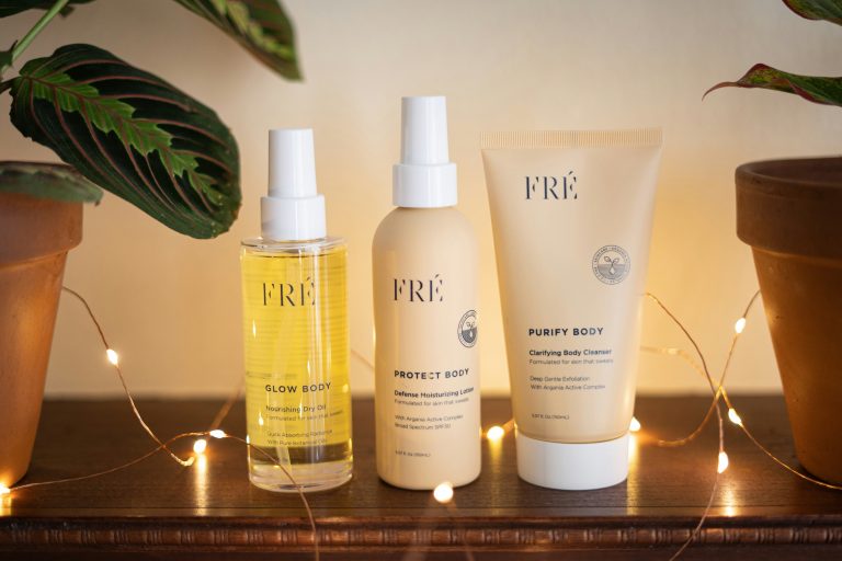 A trio of FRÉ body care products—Glow Body dry oil, Protect Body moisturizer, and Purify Body cleanser—displayed on a wooden surface with warm string lights and potted plants.