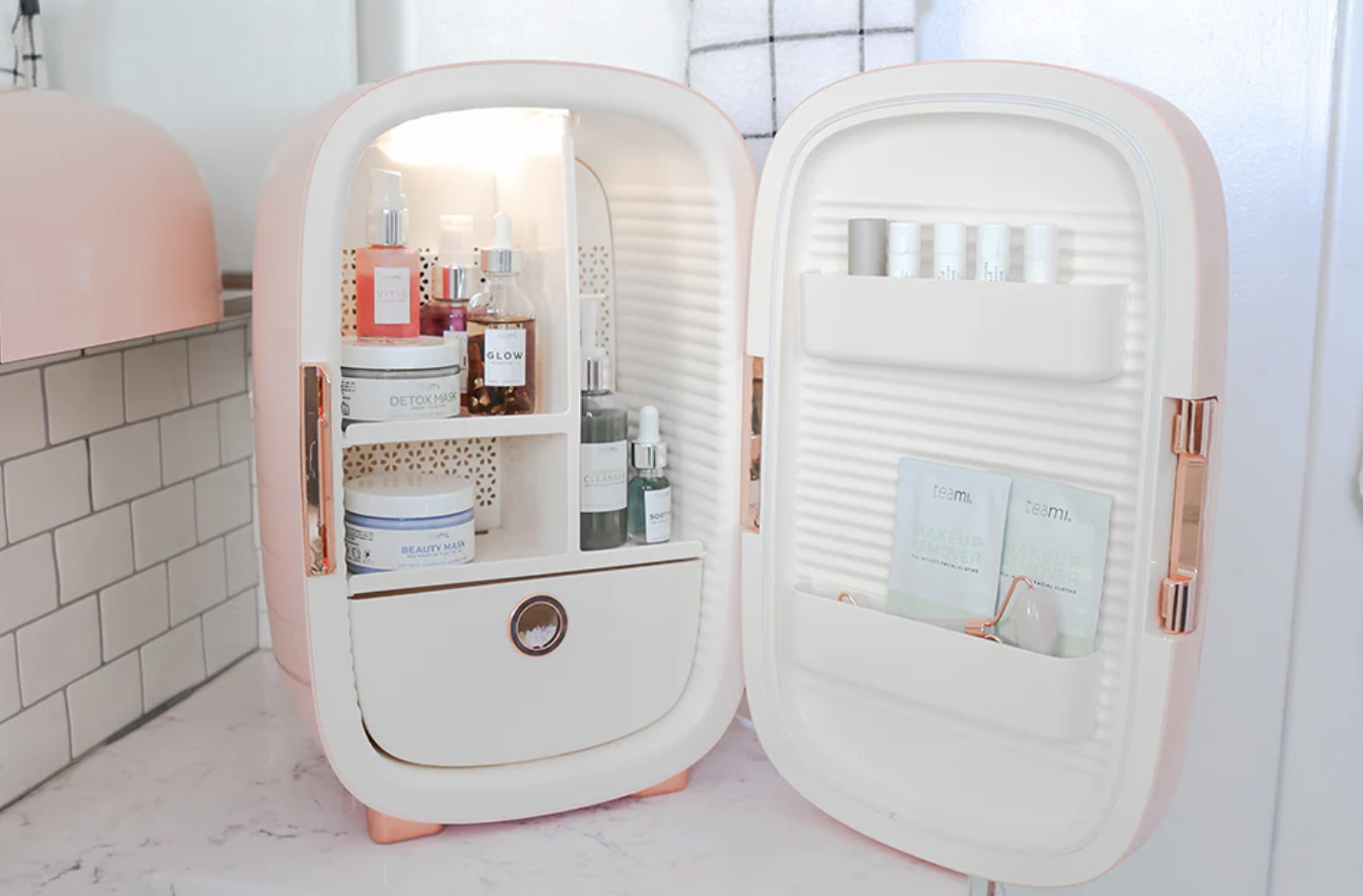 An open pink and white mini fridge neatly stocked with skincare products, serums, and masks, designed for beauty and cosmetic storage on a bathroom counter.