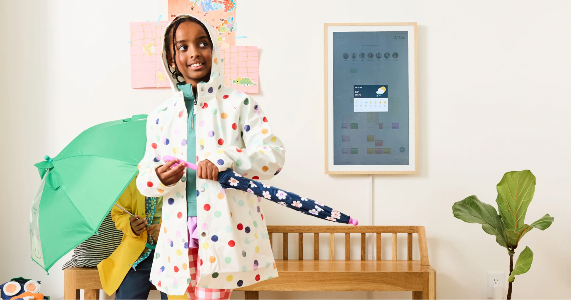 A child in a polka-dot raincoat holds an umbrella beside a Hearth Display screen showing a rainy weather forecast for family planning.