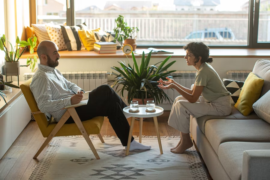 A woman speaks expressively during an in-person therapy session with a male therapist seated across from her in a cozy, sunlit room decorated with plants and soft furnishings.