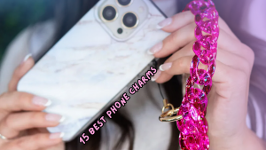 A glossy pink chain phone charm attached to a marble-patterned phone case, held by a person with a French manicure. The text "15 Best Phone Charms" is overlaid in bold pink bubble letters, highlighting the charm as a stylish and trendy accessory pick.