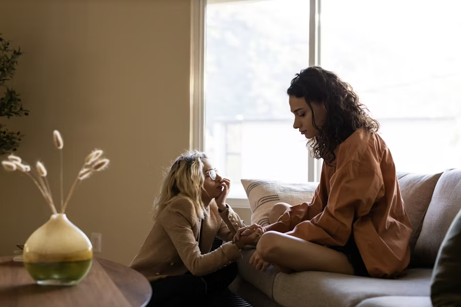 Two women sit closely on a couch in a warmly lit room, holding hands and sharing an emotional, supportive conversation, conveying empathy and care.